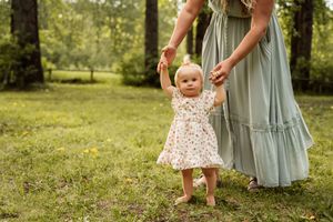 Young baby in floral dress takes first steps while holding hands with parent in blue tiered dress in sunny garden setting.