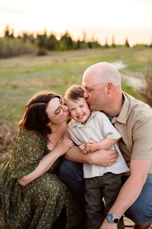 A family of three shares a warm moment at golden hour, with parents kissing their laughing toddler son on each cheek outdoors.