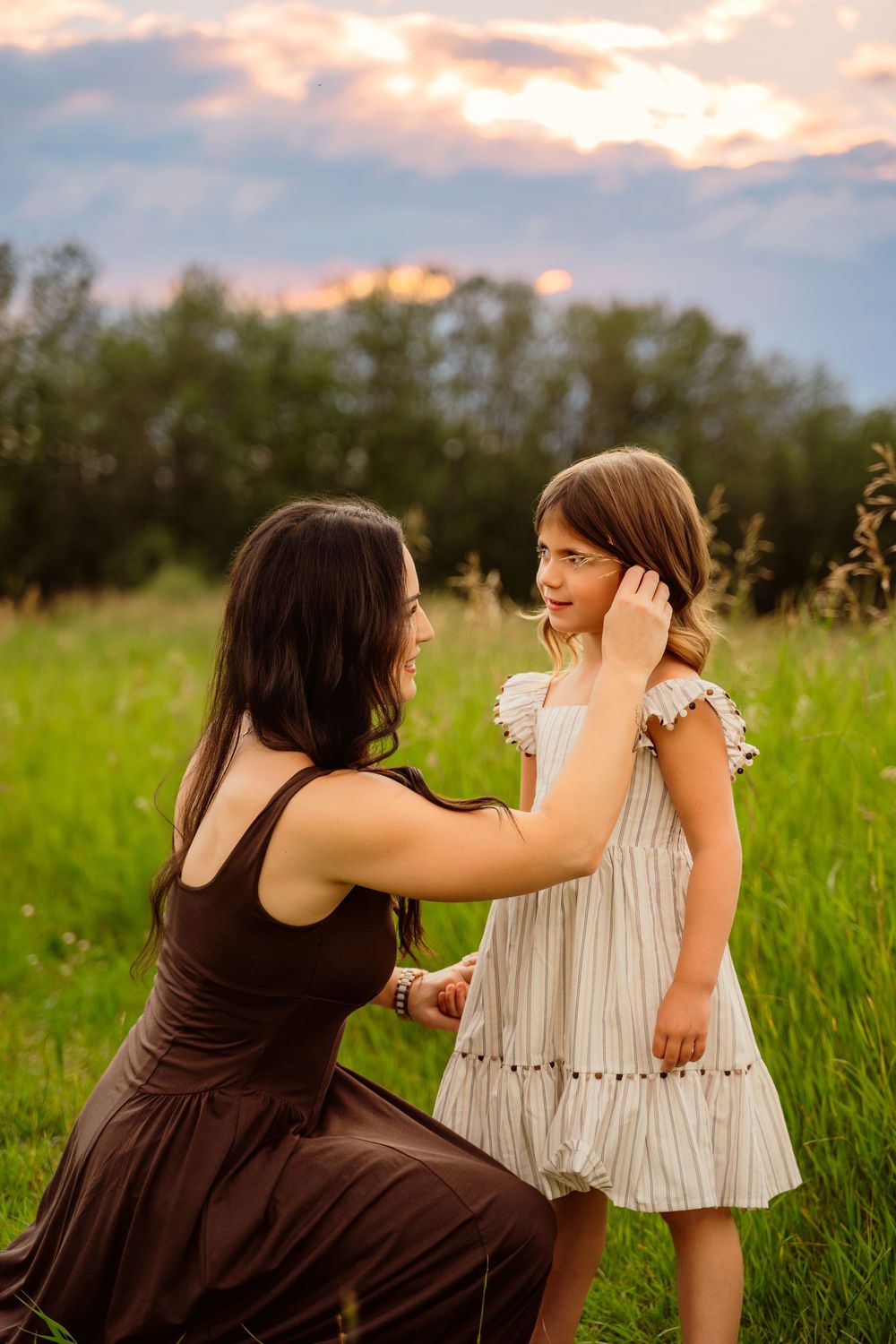 A mother kneels in a green field at sunset, gently tucking her young daughter's hair behind her ear.