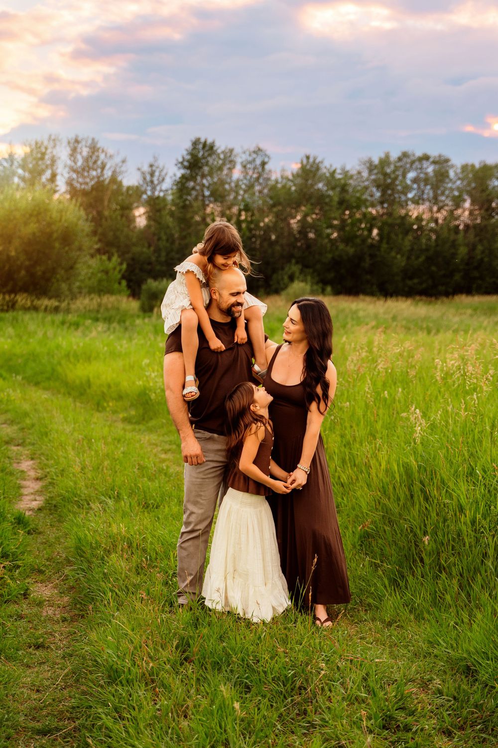 Family of four poses in a lush green field at sunset, with two daughters and parents in elegant brown and white outfits.