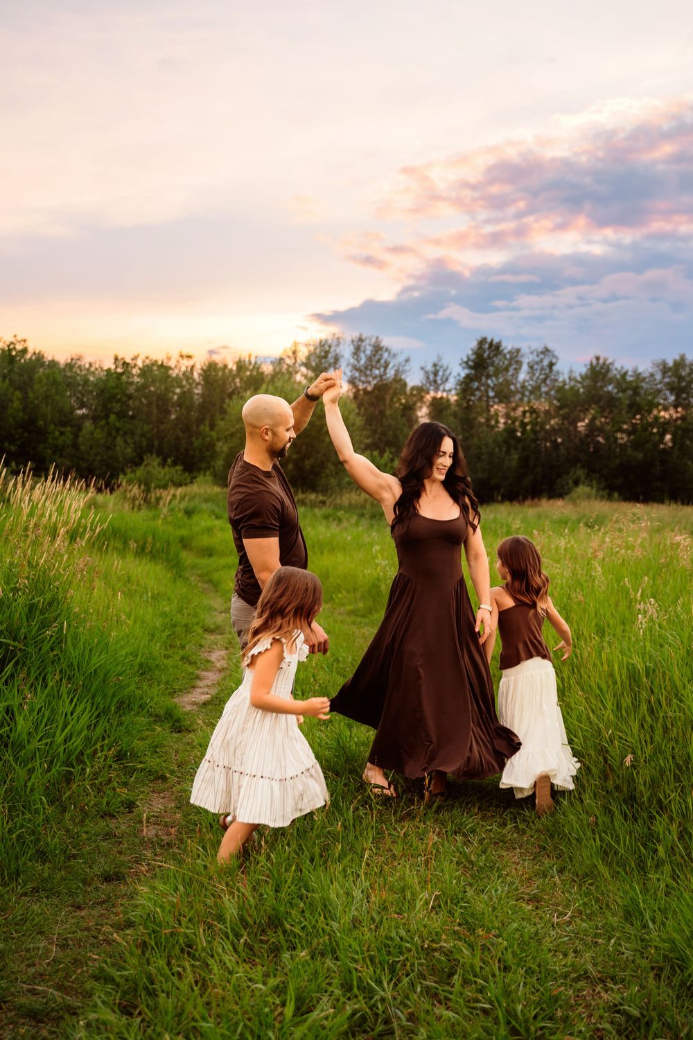 Family dancing in a green field at sunset, parents spinning two daughters in white dresses on a scenic nature path.