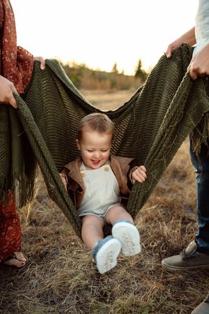 A knitted green blanket being held up while a small child sits underneath in a grassy field at sunset.