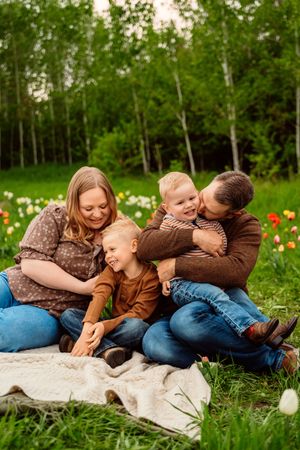 Family of four sitting on blanket in flower garden, parents hugging laughing children outdoors.