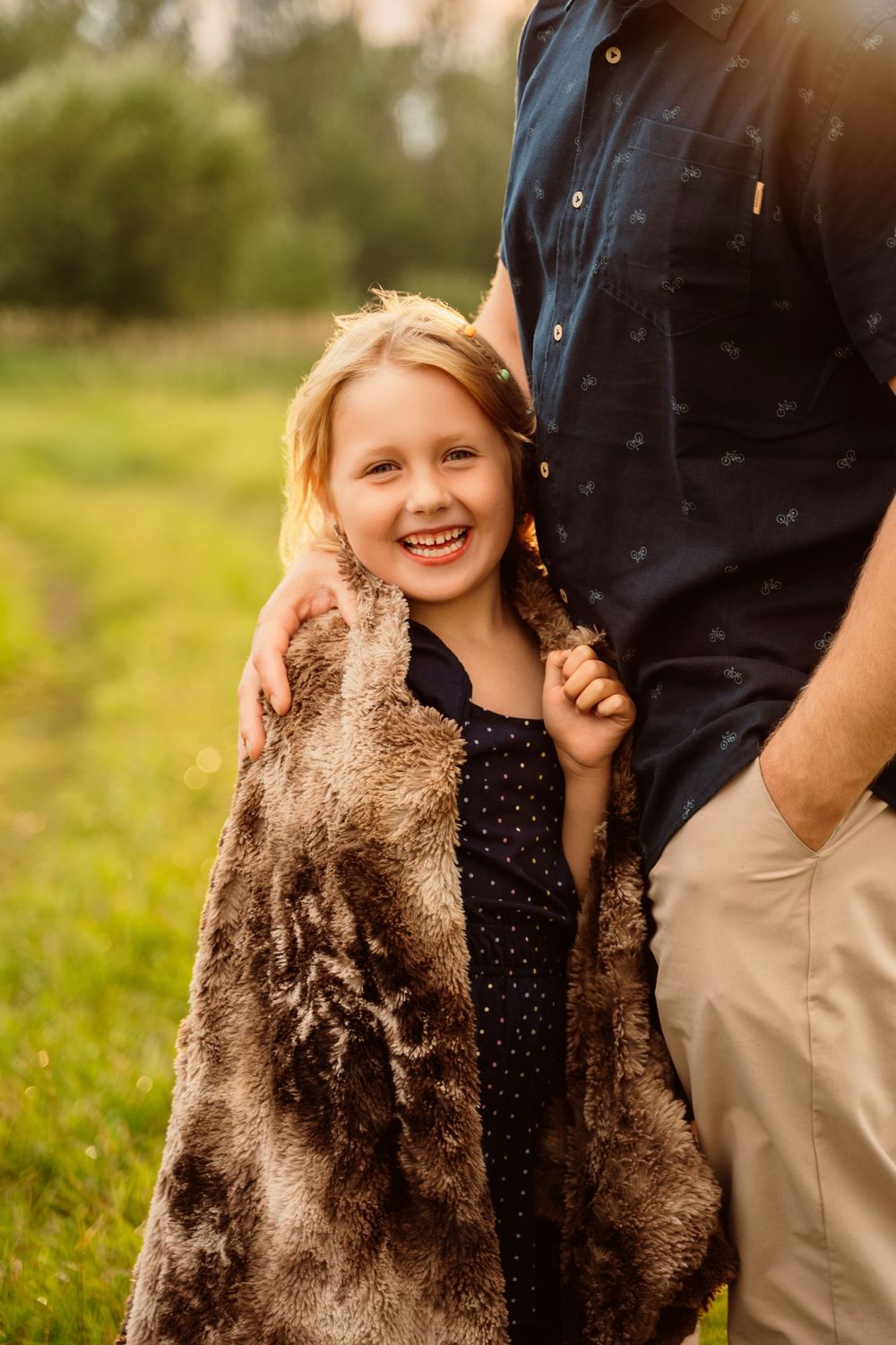 A smiling blonde girl wrapped in a fur blanket stands beside a man in a navy shirt in a sunny green field.