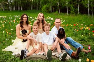 Family of six poses with a puppy in a colorful tulip field surrounded by green trees on a sunny day.