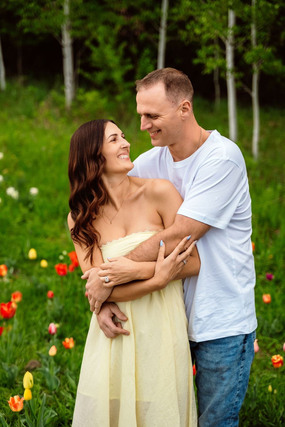 A smiling couple embraces in a colorful tulip field with green trees, woman in yellow dress, man in white tee and jeans.