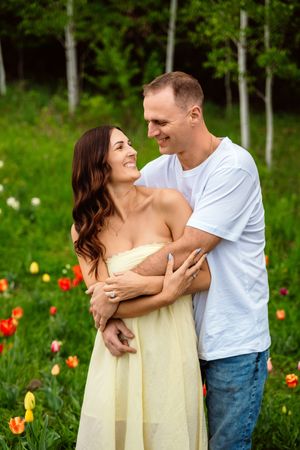 A smiling couple embraces in a colorful tulip field with green trees, woman in yellow dress, man in white tee and jeans.