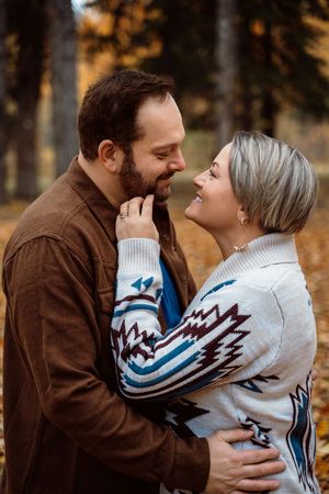 A couple in a brown jacket and white patterned sweater share an intimate moment in an autumn forest setting.