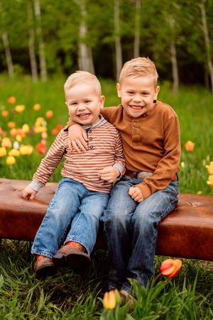 Two smiling brothers in brown tops and jeans sit on a bench surrounded by colorful tulips and green trees.