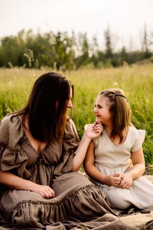 Two people in flowing dresses share a joyful moment while sitting together in a sunlit meadow during golden hour.