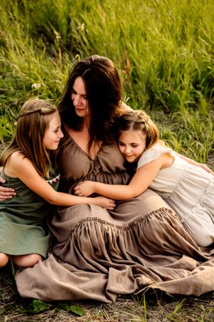 Three figures share a tender embrace while sitting together in tall grass on a sunny day, wearing flowing dresses.