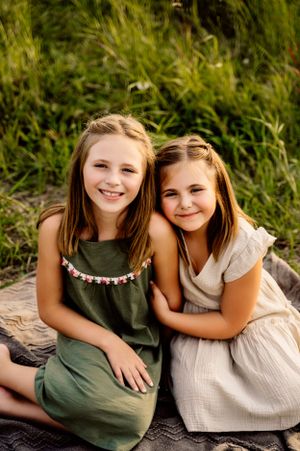 Two young people in summer dresses sit together outdoors on a blanket with green foliage in the background.