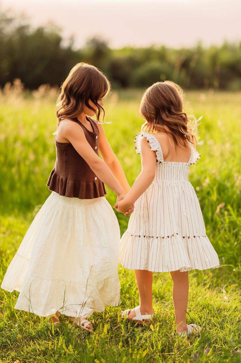 Two young girls in white dresses hold hands in a sunny green meadow, viewed from behind.