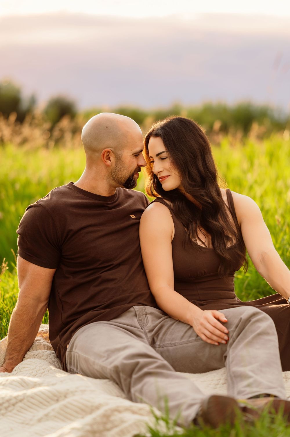 A couple sits intimately on a blanket in a sunlit green field, nearly touching foreheads in a romantic moment.