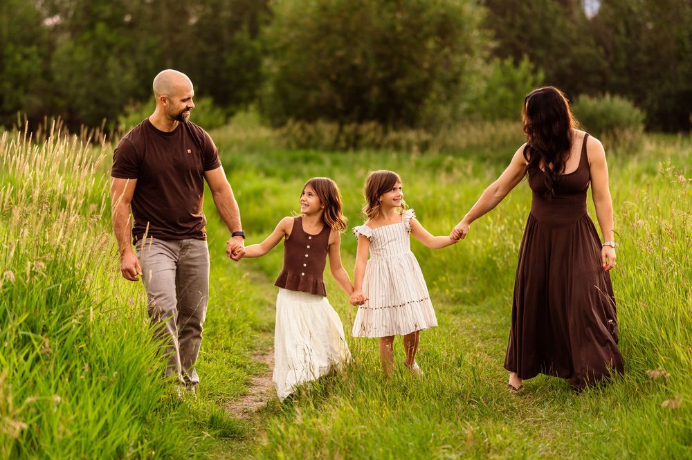 Family of four walking hand-in-hand through a lush green meadow path, parents with two young daughters in summer dresses.
