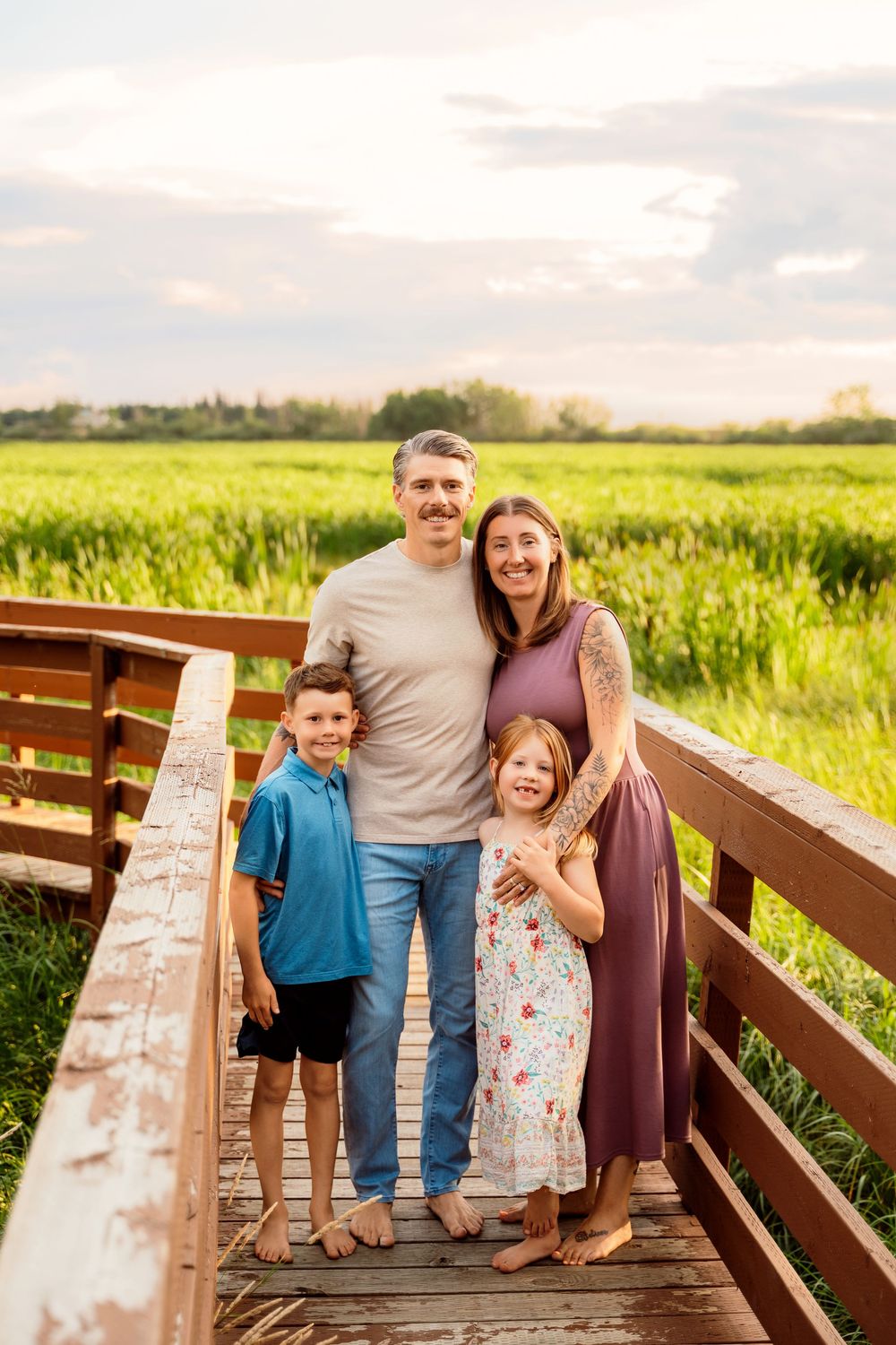 Family of four smiling on a wooden bridge surrounded by lush green fields during golden hour.