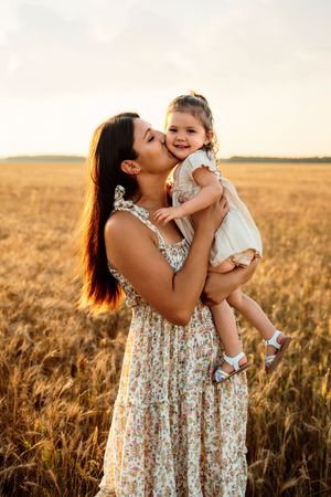 Parent and child share a tender moment in matching floral dresses while standing in a golden wheat field at sunset.