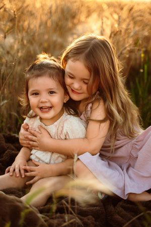 Two young children share a joyful embrace while sitting together in a golden-lit field at sunset.