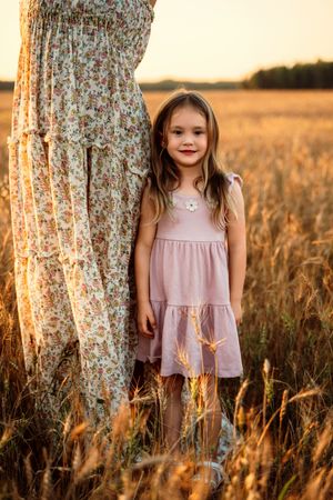 A child in a pink dress stands beside a tree trunk in a golden wheat field at sunset.