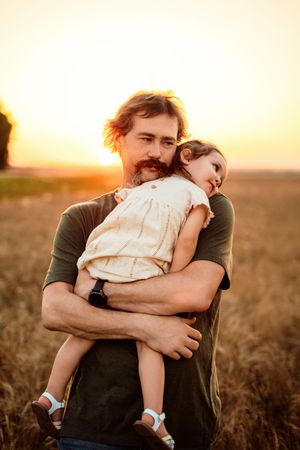 A tender sunset moment in a golden field as a figure holds a small child wearing a white dress.
