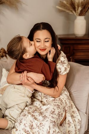 A young child lovingly kisses their smiling mother on the cheek while sitting together on a cozy sofa.