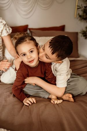Two young siblings sharing a sweet moment on a bed, with the older boy kissing his little sister on the cheek.