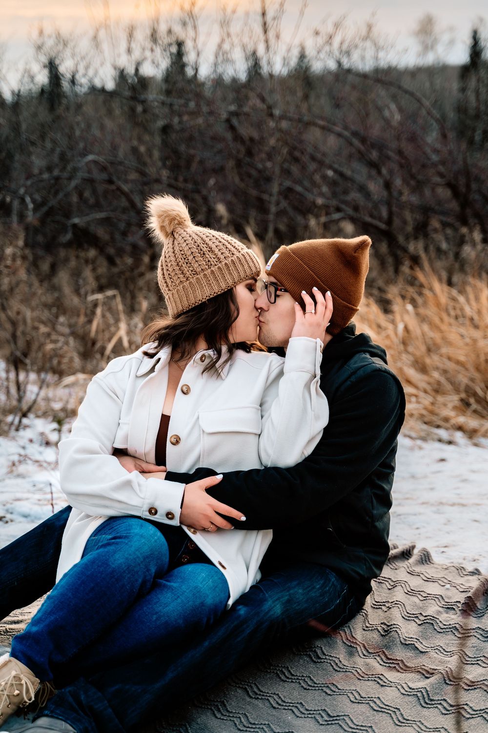 A couple in winter clothing sharing an intimate moment on a blanket in a snowy outdoor setting at dusk.