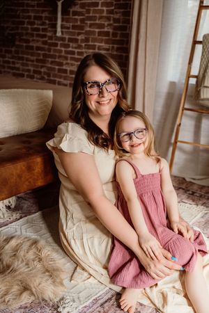 A smiling woman and young girl in glasses sit together on a rug in a cozy boho-style room.