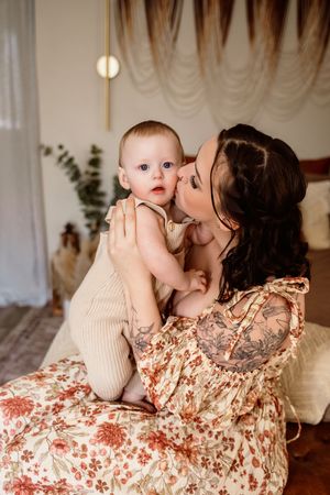 A tattooed mother in a floral dress kisses her blue-eyed baby in a cozy bedroom setting.
