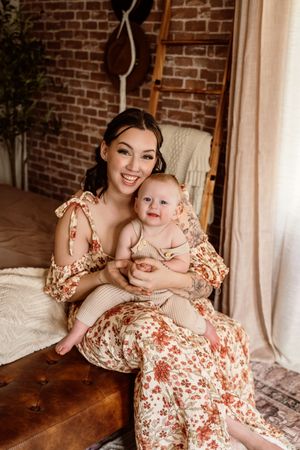Smiling woman in floral dress holding happy baby in boho bedroom with brick wall backdrop.