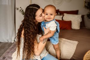 A young mother with long curly hair kisses her smiling baby dressed in denim overalls in a cozy bedroom.