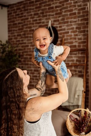 Tattooed mother joyfully lifting laughing baby in denim overalls in a cozy brick-walled room.