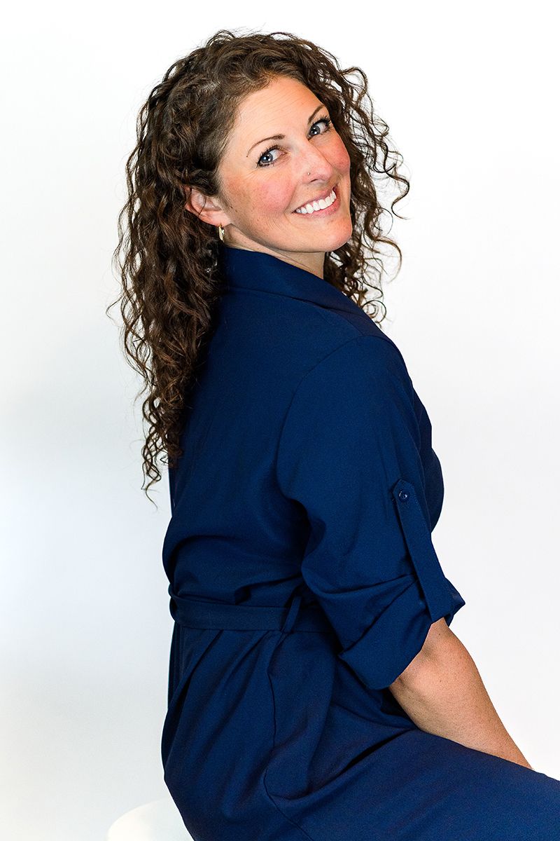A smiling woman with curly brown hair poses in a navy blue dress against a white studio background.