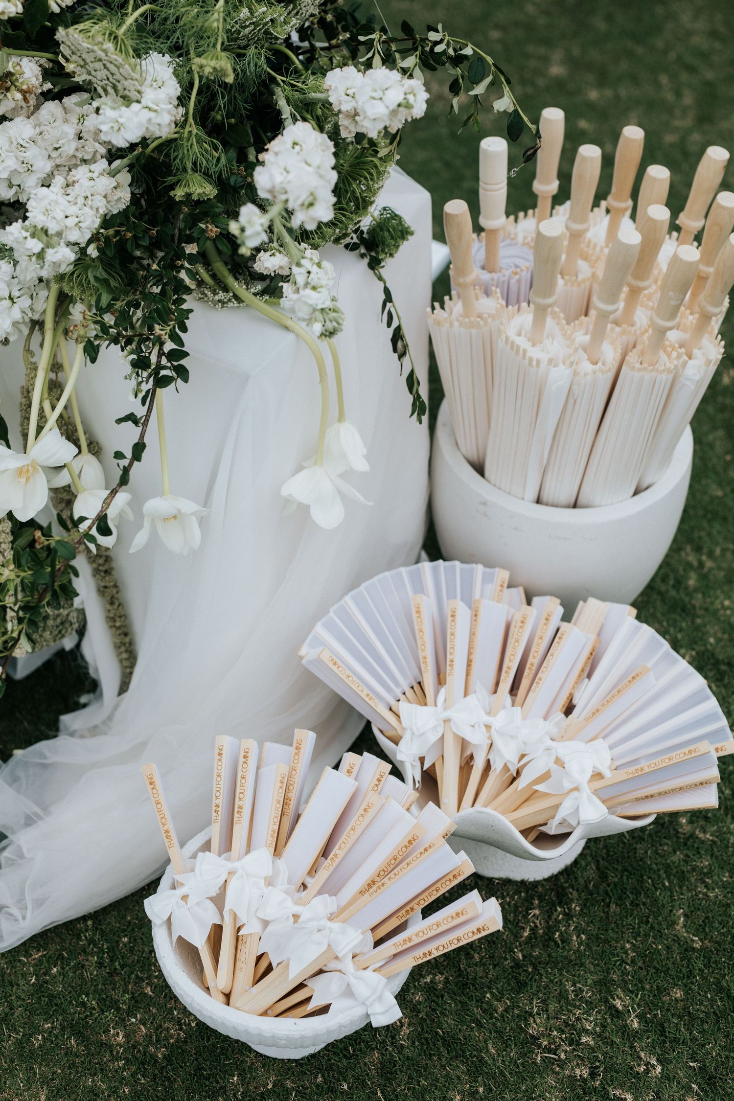 White paper hand fans and parasols displayed in white buckets at outdoor wedding ceremony.