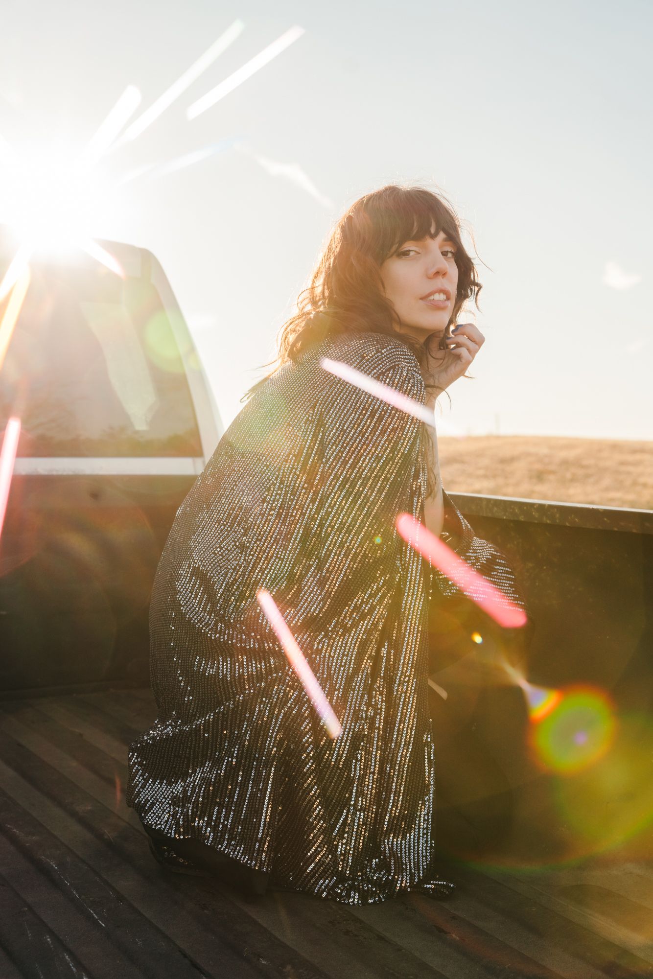 Woman with dark bangs crouches on a wooden truck bed in a sparkling sequin dress, bathed in golden sunset light and lens flare.