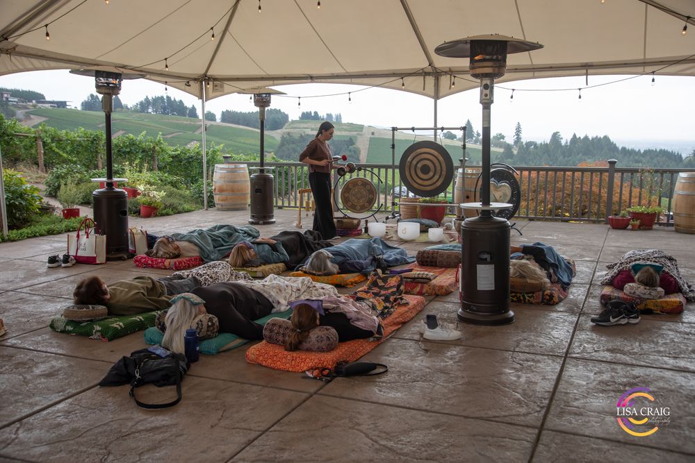 People relax on mats and blankets during an outdoor sound bath session under a covered patio with gongs and vineyard views.