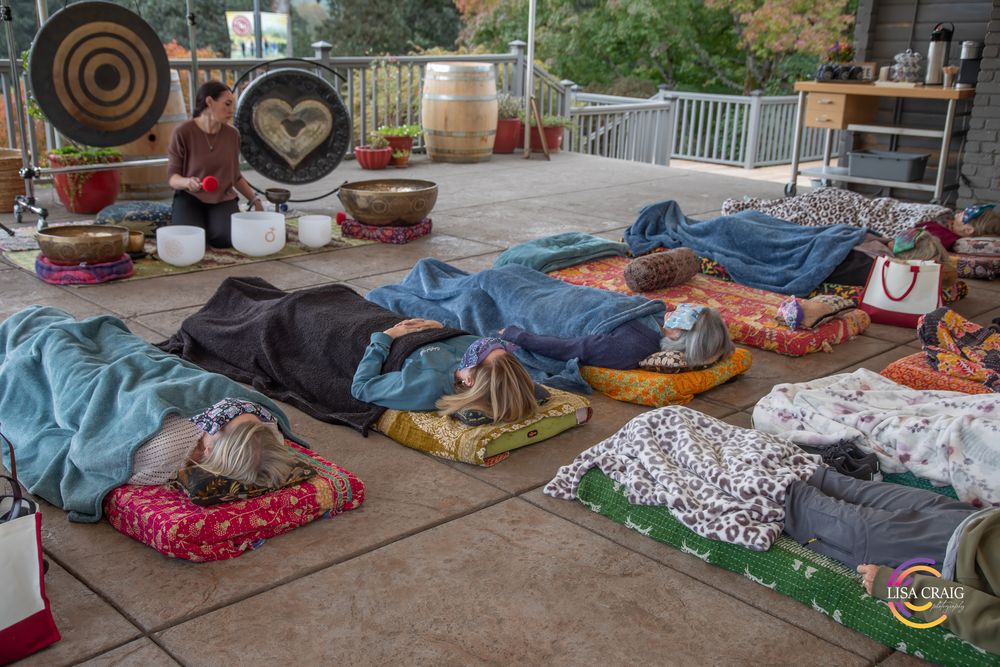 People lie on colorful mats and blankets on an outdoor patio during a sound bath meditation while a facilitator plays singing bowls nearby.