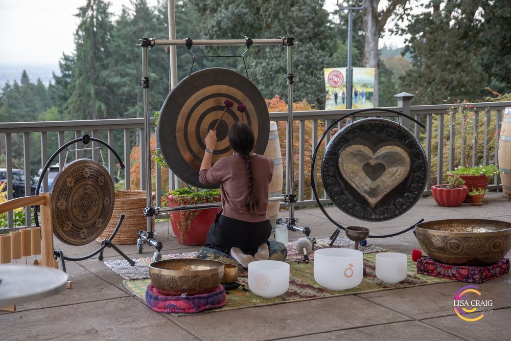 Person kneeling on a rug plays a large gong with a heart design, surrounded by singing bowls on an outdoor deck with forest views.