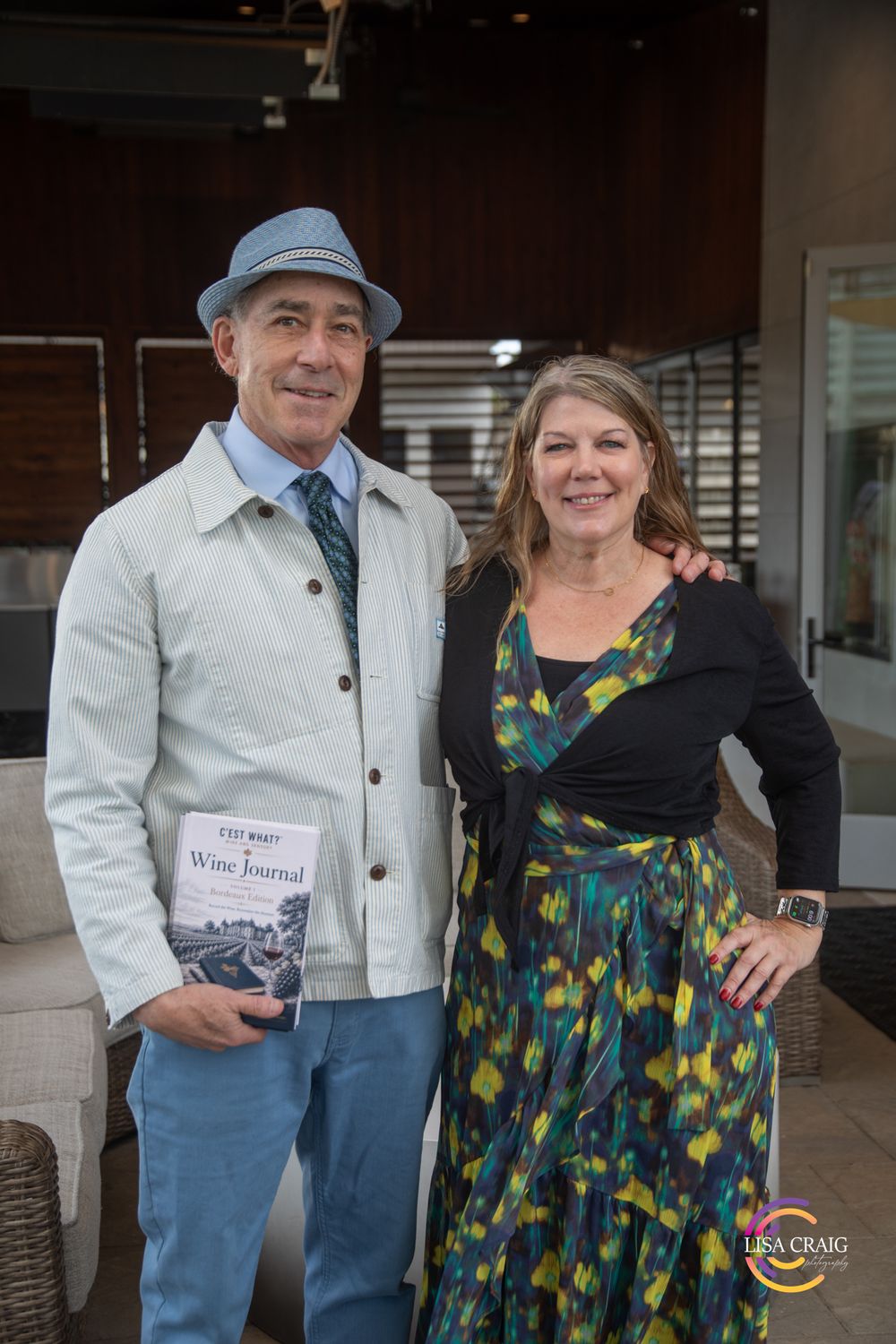 A smiling man in a blue hat holding a Wine Journal poses with a smiling woman in a colorful floral dress at an indoor event.