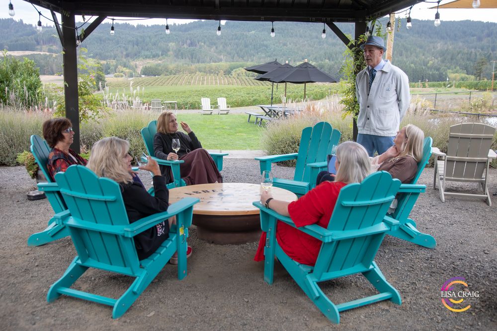 Guests relax in turquoise Adirondack chairs around a table on a vineyard patio while a host speaks, with rolling hills in the background.