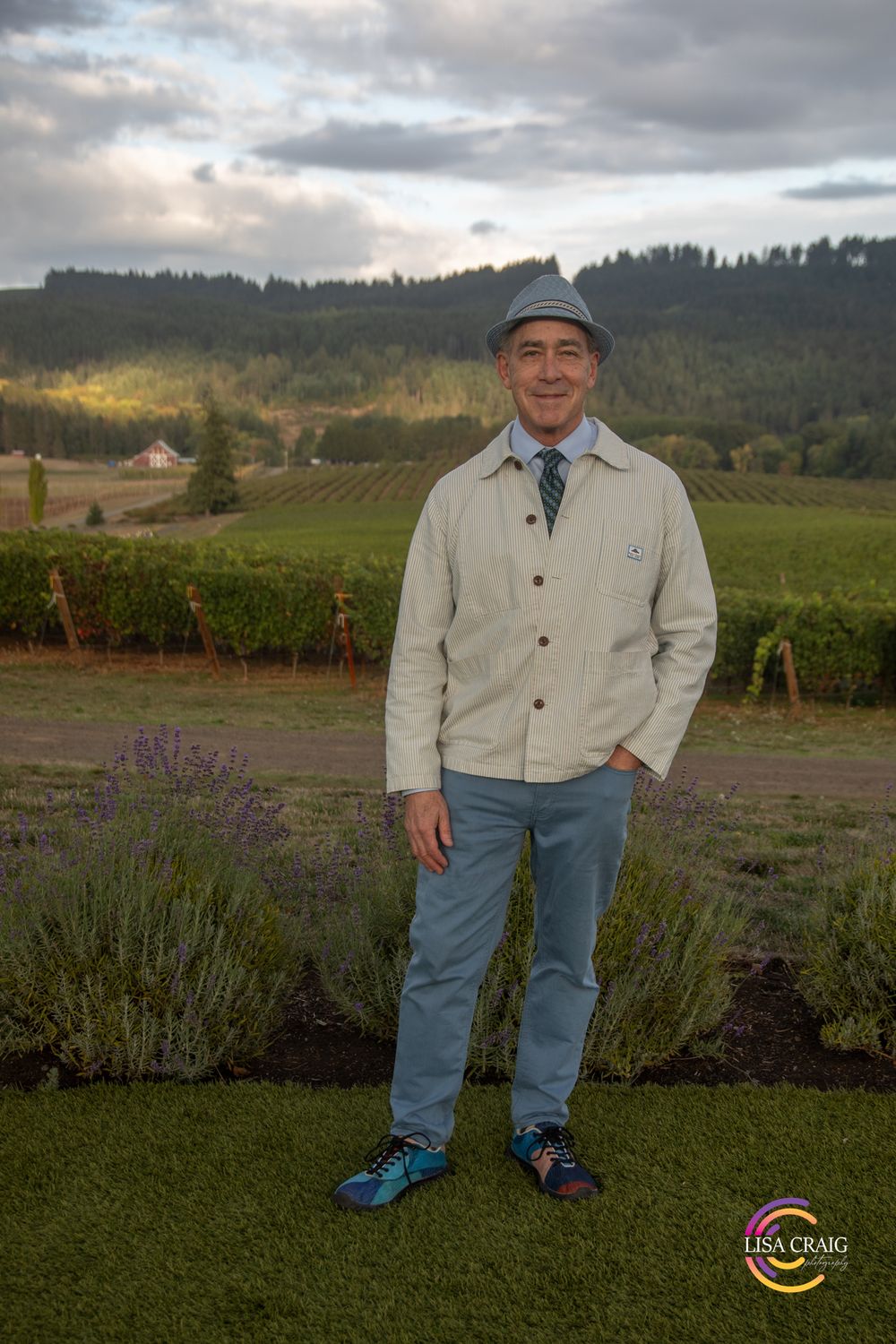 Man in a gray hat, cream jacket and blue jeans stands smiling beside lavender with rolling vineyard hills and cloudy sky behind him.