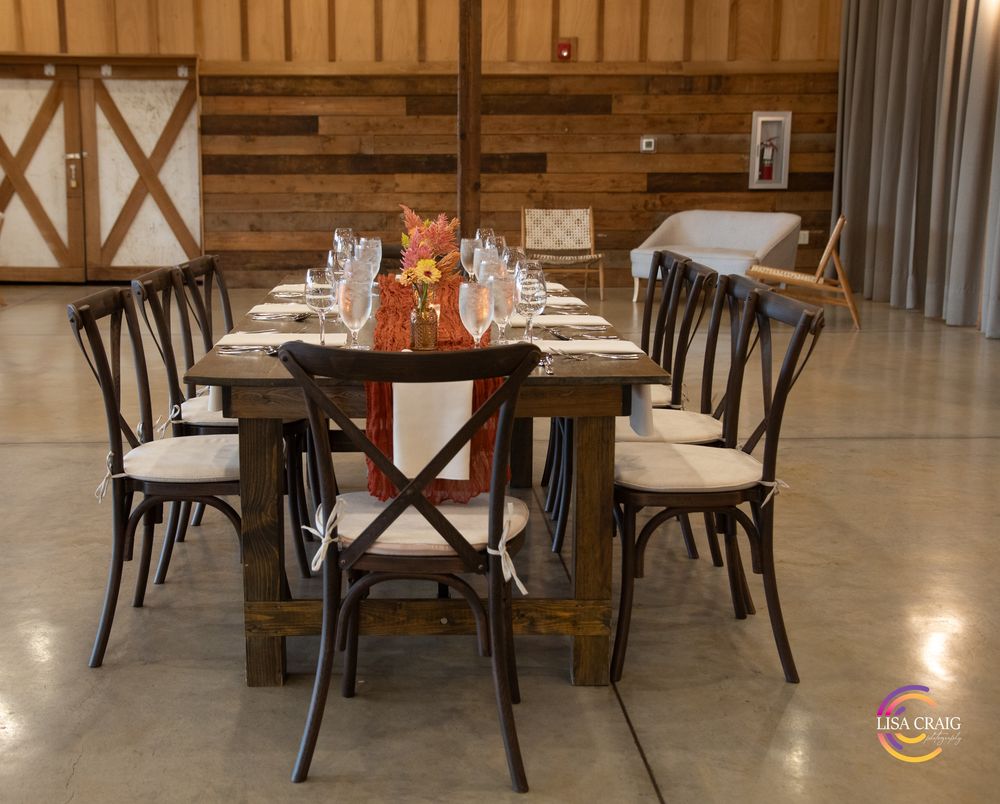Rustic wooden farm table with orange floral centerpiece, wine glasses and crossback chairs in a barn wedding venue with sliding doors.