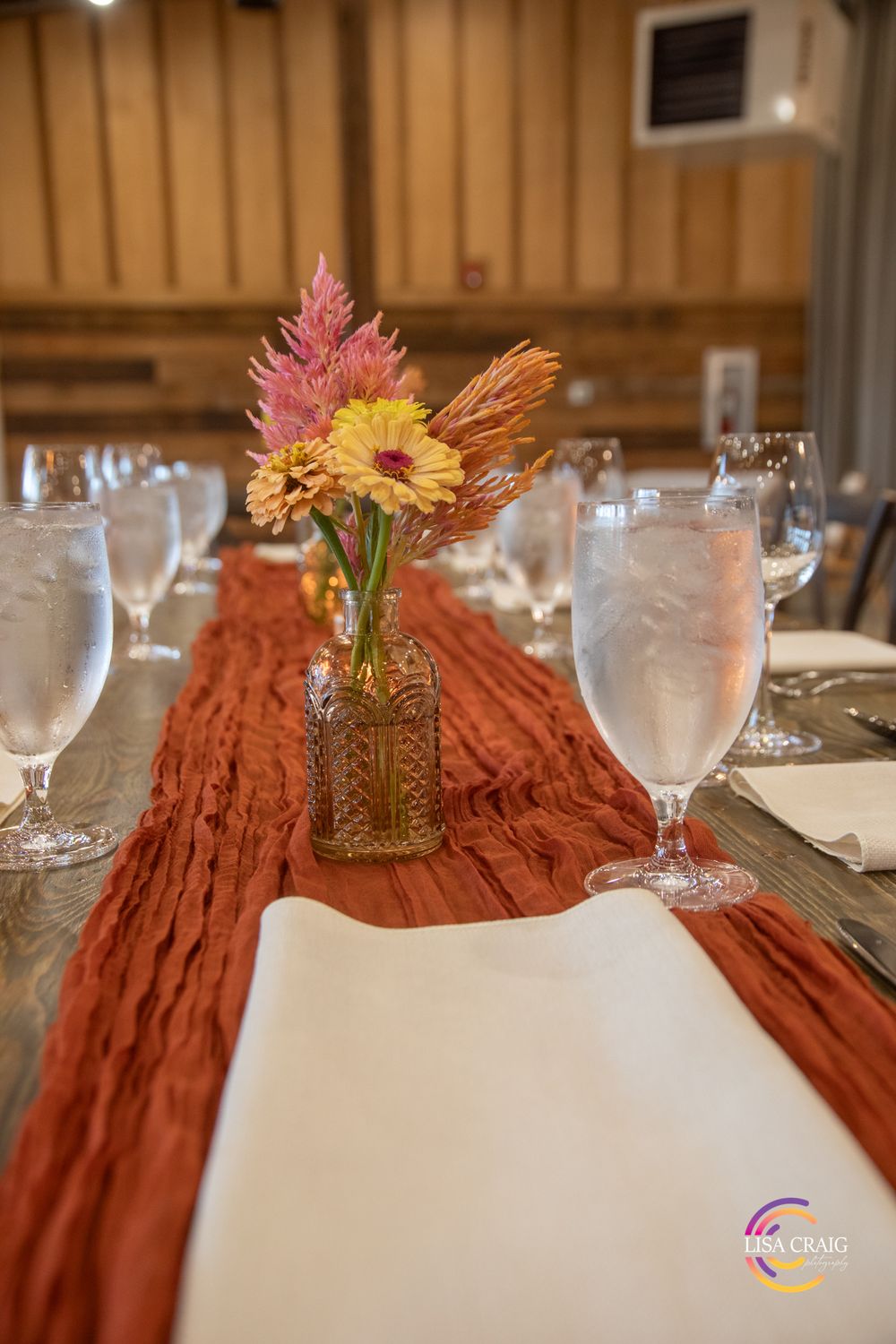 Rustic wedding table with a terracotta gauze runner, water glasses, cream napkins, and a vintage glass bud vase of pink and yellow flowers.