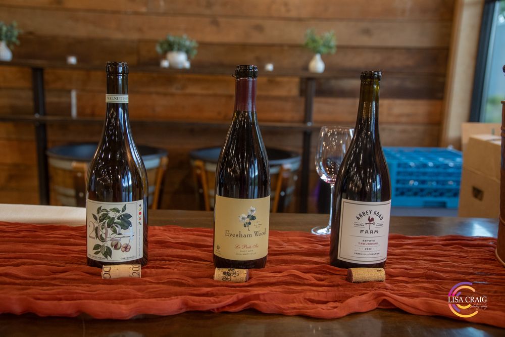 Three bottles of red wine with labels on a red table runner at a rustic tasting room, with an empty wine glass nearby.