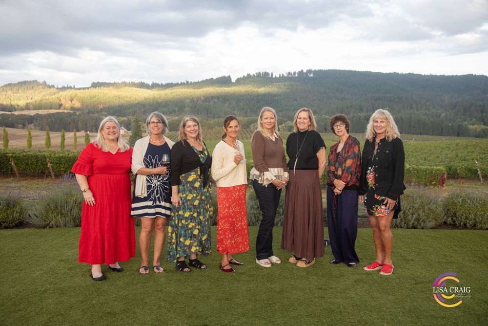 Group of nine women in colorful dresses pose together on a green lawn overlooking vineyard hills at sunset.