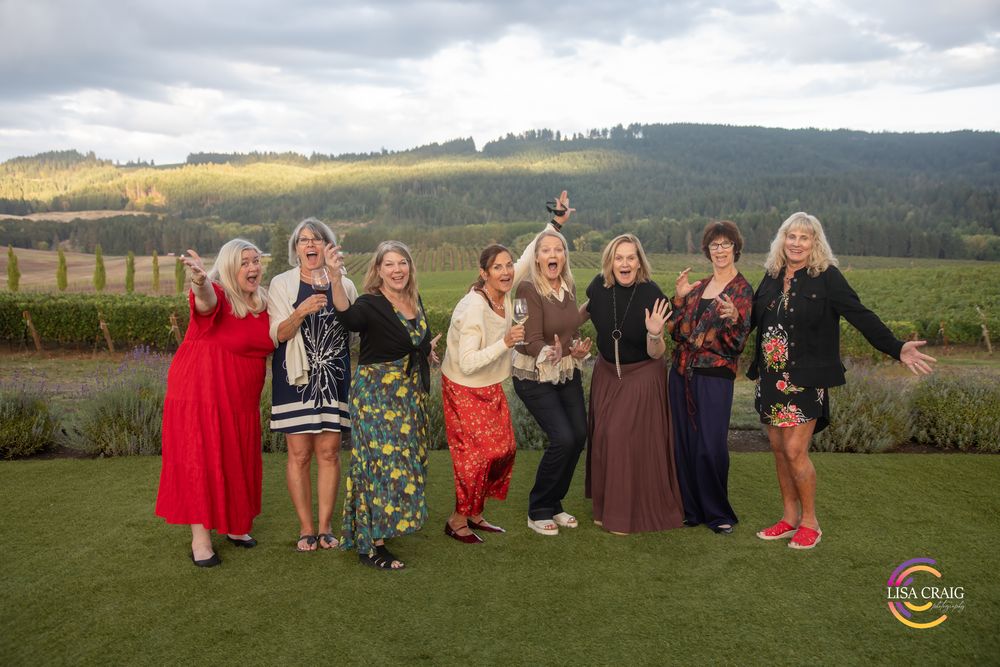 Group of nine women in colorful dresses laughing and posing together on a green lawn with vineyard rows and rolling hills behind them.