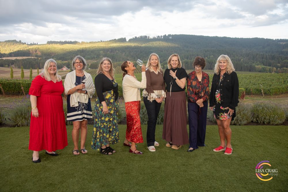 Eight women in colorful dresses and casual outfits pose together on a green lawn with rolling vineyard hills in the background.