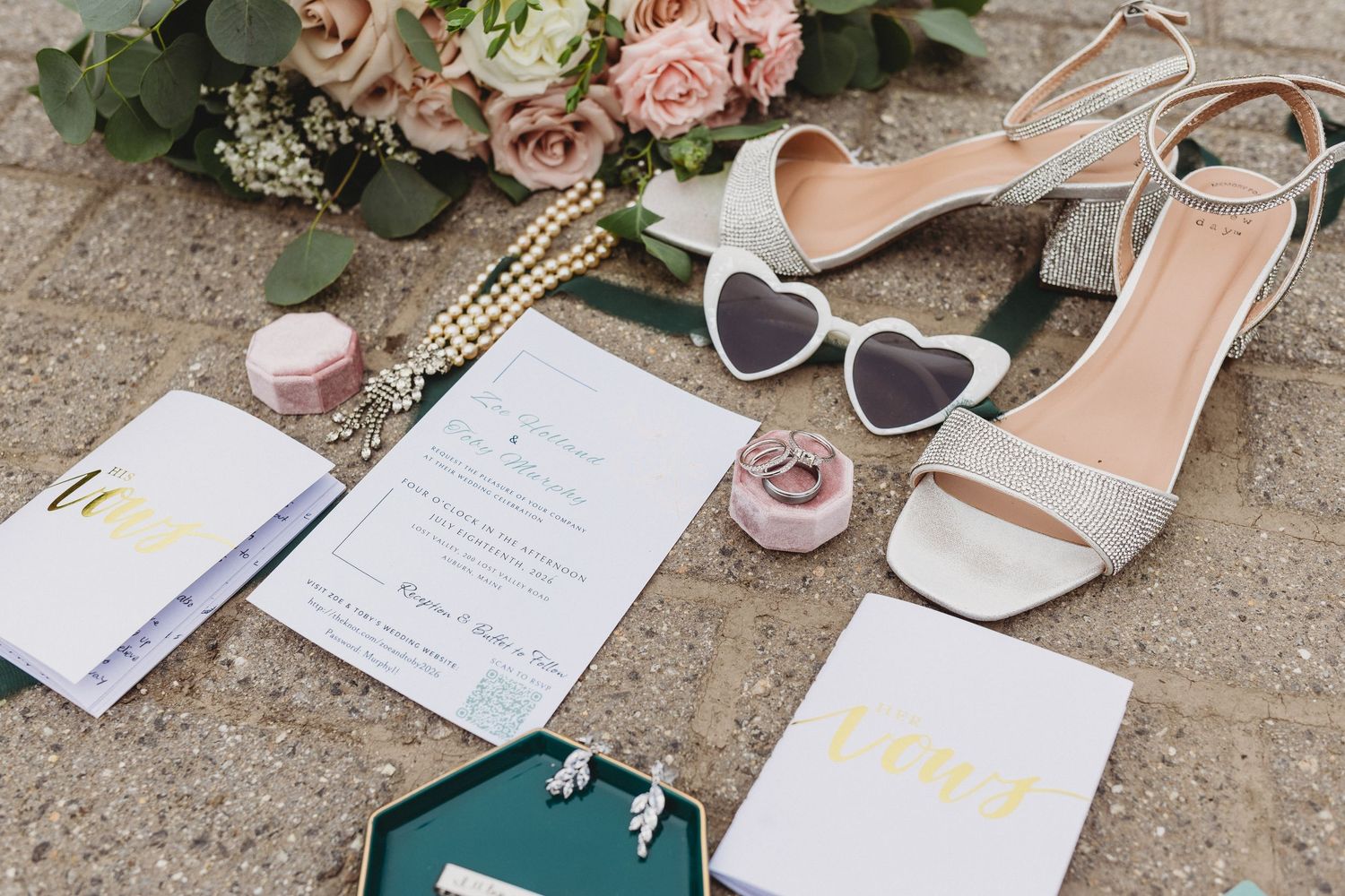 Wedding stationery arranged with silver bridal heels, heart-shaped sunglasses, jewelry, ring boxes, and pink flowers.
