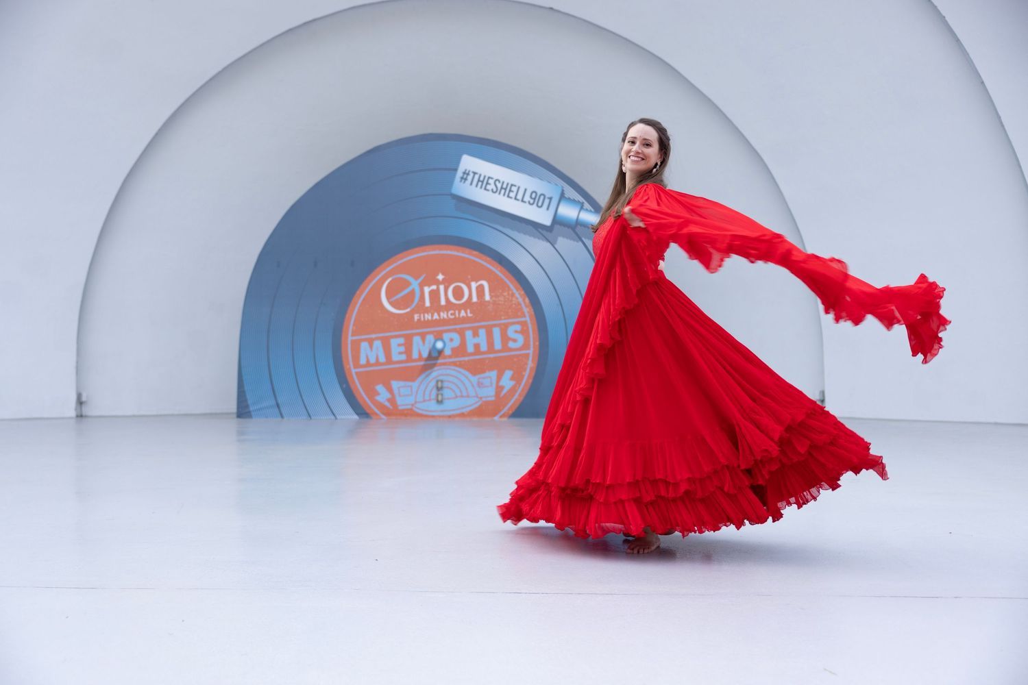 Woman in a flowing red ruffled dress twirls on a white stage in front of a large Orion Memphis vinyl record sign.