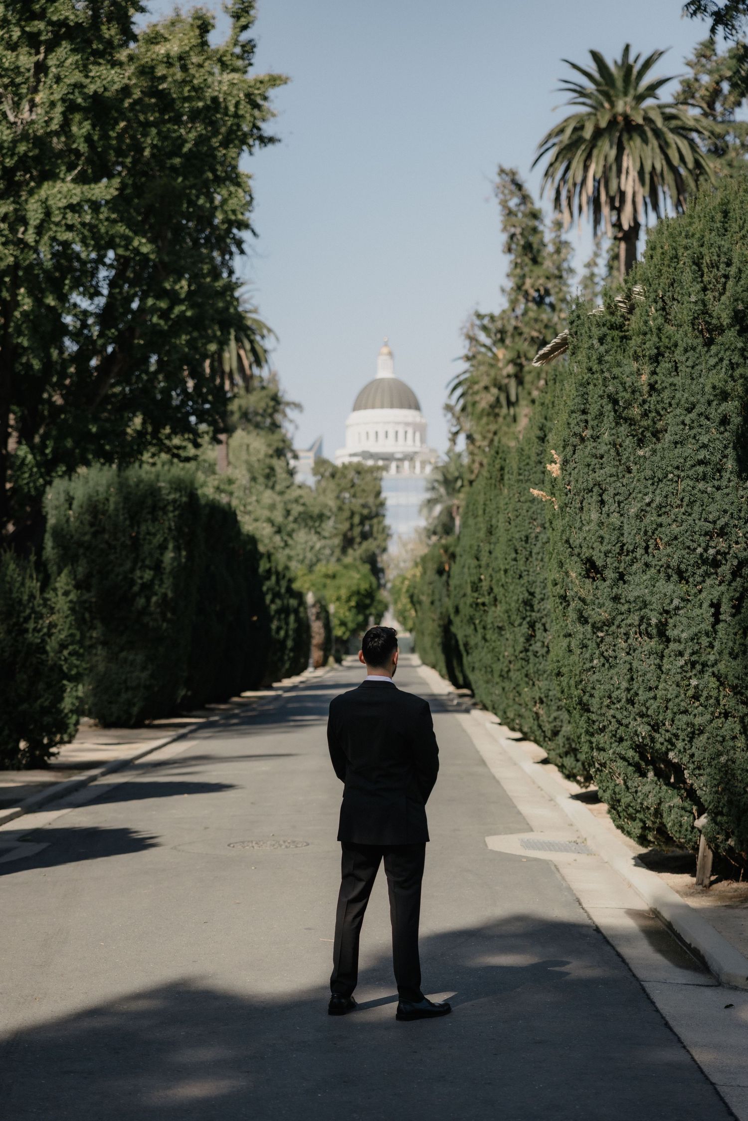 Man in a dark suit stands on a tree-lined street, facing a distant domed capitol building under a clear blue sky.